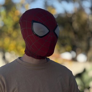 Puede incluir: Una m&aacute;scara de Spider-Man roja y negra con cubiertas oculares de malla blanca. La m&aacute;scara tiene un patr&oacute;n de telara&ntilde;a y cubre toda la cabeza y el cuello. La persona lleva una camiseta de manga larga beige.
