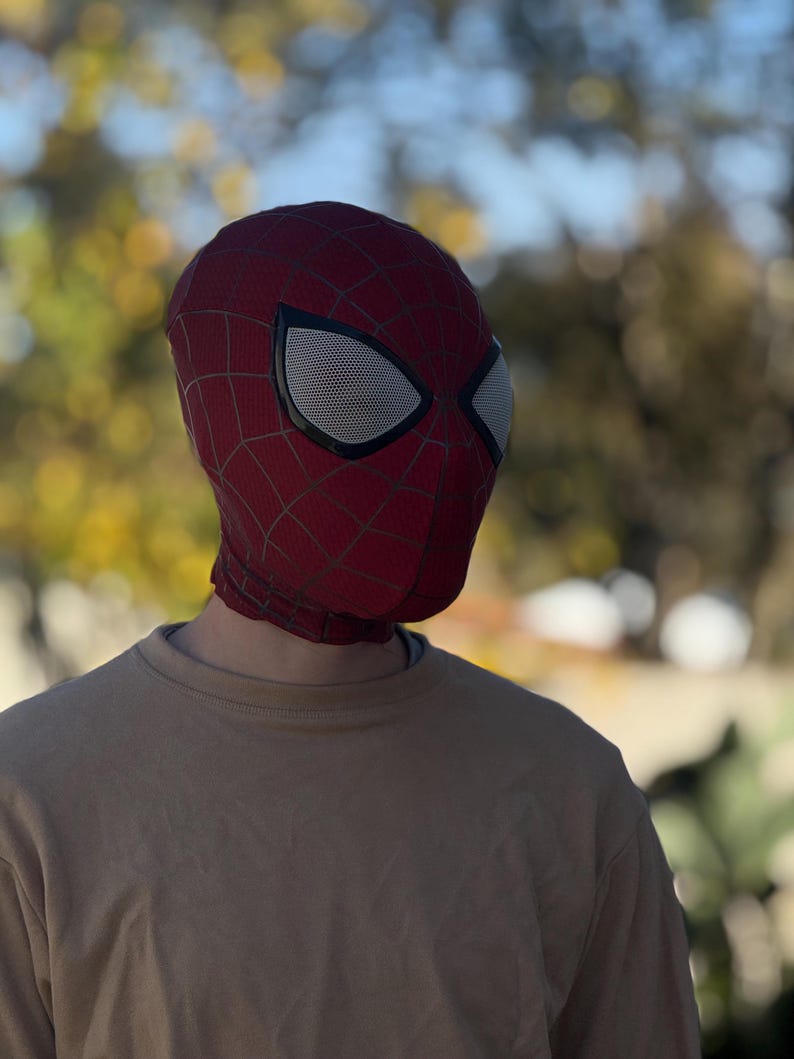 Puede incluir: Una m&aacute;scara de Spider-Man roja y negra con cubiertas oculares de malla blanca. La m&aacute;scara tiene un patr&oacute;n de telara&ntilde;a y cubre toda la cabeza y el cuello. La persona lleva una camiseta de manga larga beige.