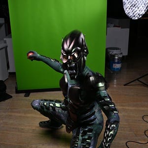 May include: A Green Goblin costume with a metallic helmet and armor. The costume features a detailed design with a menacing mask and sharp teeth. The character is in a crouching position, holding a red object.