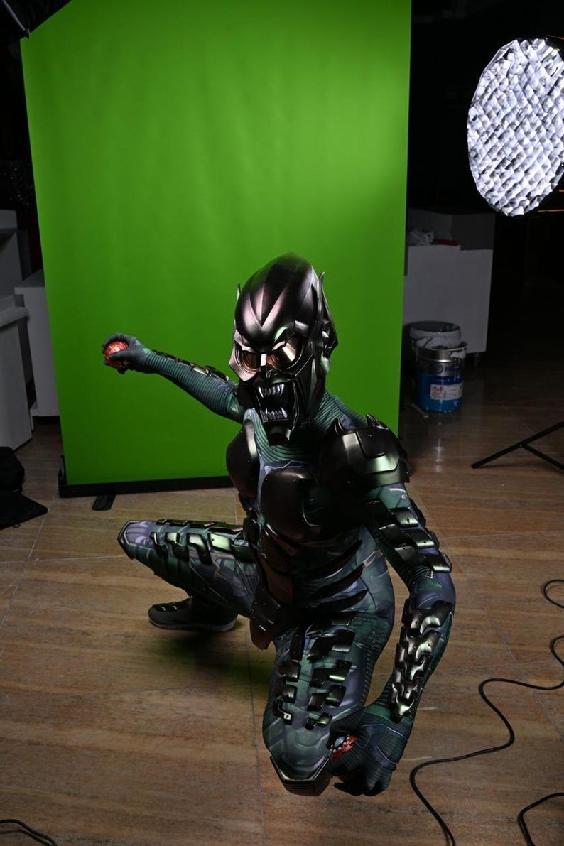 May include: A Green Goblin costume with a metallic helmet and armor. The costume features a detailed design with a menacing mask and sharp teeth. The character is in a crouching position, holding a red object.