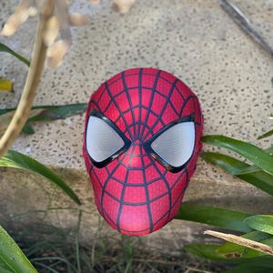 Może przedstawiać: Czerwono-czarna maska Spider-Mana z białymi siatkowymi osłonami oczu. Maska ma wz&oacute;r pajęczyny i spoczywa na teksturowanej powierzchni, otoczona zielenią.