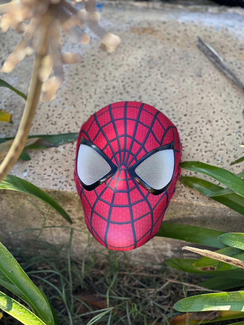 Może przedstawiać: Czerwono-czarna maska Spider-Mana z białymi siatkowymi osłonami oczu. Maska ma wz&oacute;r pajęczyny i spoczywa na teksturowanej powierzchni, otoczona zielenią.