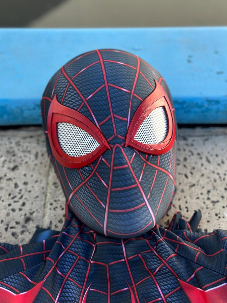 May include: A black and red Spider-Man mask and costume. The mask has red eye frames with white mesh eye coverings. Red lines form a web pattern across the black surface of the mask and costume.
