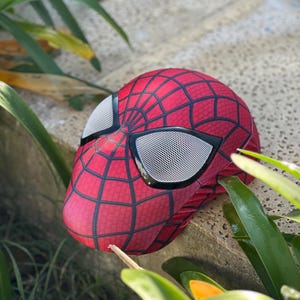 Może przedstawiać: Czerwono-czarna maska Spider-Mana z wzorem pajęczyny i siatkowymi osłonami oczu. Maska spoczywa na kamiennej powierzchni, otoczona zielonymi liśćmi. Maska jest repliką nakrycia głowy superbohatera.