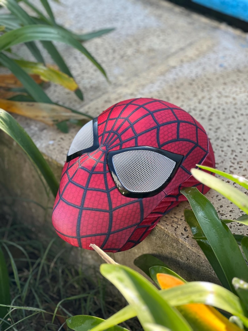 Może przedstawiać: Czerwono-czarna maska Spider-Mana z wzorem pajęczyny i siatkowymi osłonami oczu. Maska spoczywa na kamiennej powierzchni, otoczona zielonymi liśćmi. Maska jest repliką nakrycia głowy superbohatera.