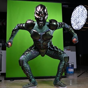 May include: A Green Goblin costume with a metallic mask and armor. The costume features a green and black color scheme with intricate detailing. The character is posed in a crouching position, holding a small red object in each hand.