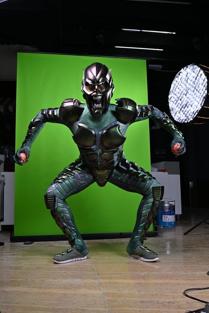 May include: A Green Goblin costume with a metallic mask and armor. The costume features a green and black color scheme with intricate detailing. The character is posed in a crouching position, holding a small red object in each hand.