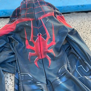 May include: A black and blue superhero costume with a red spider emblem on the back. The costume has a hood and a web-like pattern in red. The suit is made of a flexible material.