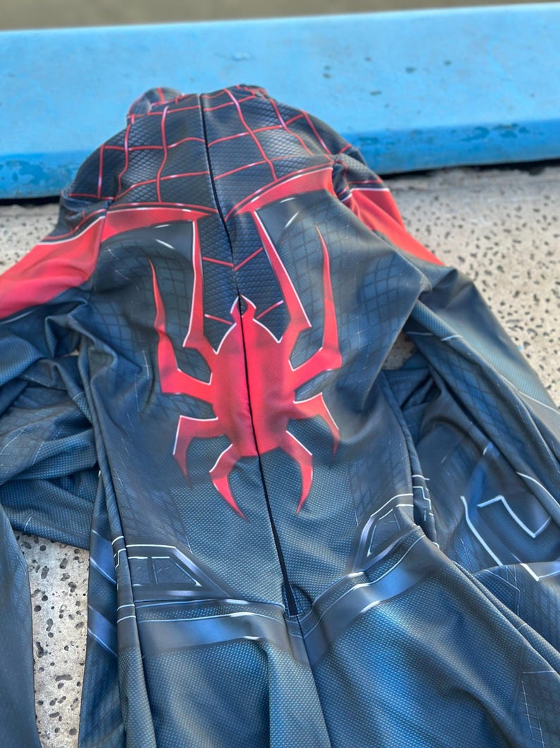 May include: A black and blue superhero costume with a red spider emblem on the back. The costume has a hood and a web-like pattern in red. The suit is made of a flexible material.