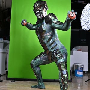 May include: A Green Goblin costume with a detailed mask and armor-like accents. The costume features a green and black color scheme, with intricate patterns and designs. The character is holding two orange and red devices.
