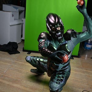 May include: A Green Goblin costume with a metallic helmet and armor, holding two pumpkin bombs. The costume features a green and black color scheme with intricate detailing. The character is kneeling on a wooden floor.