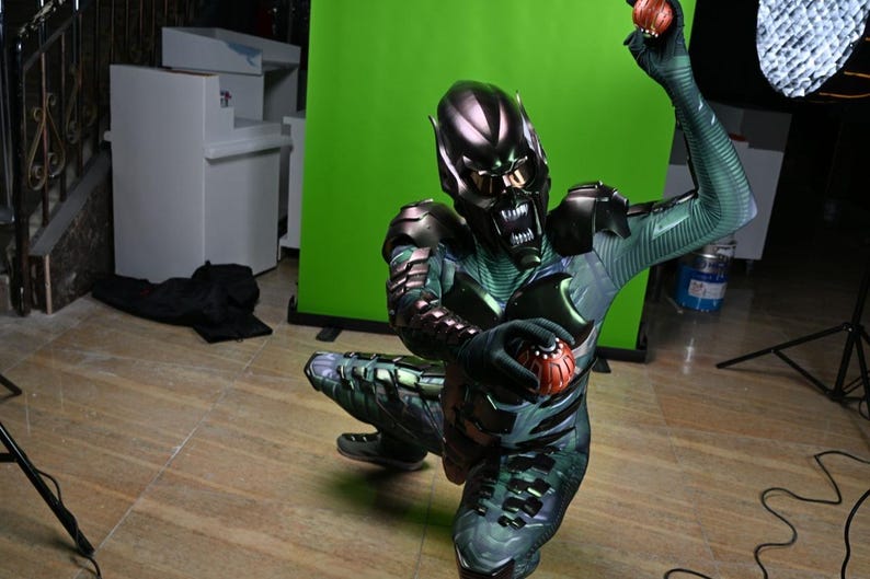 May include: A Green Goblin costume with a metallic helmet and armor, holding two pumpkin bombs. The costume features a green and black color scheme with intricate detailing. The character is kneeling on a wooden floor.