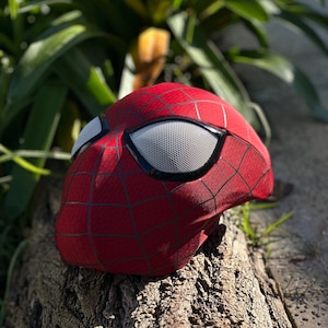 Puede incluir: Una m&aacute;scara roja de Spider-Man con telara&ntilde;as negras y lentes de malla blanca. La m&aacute;scara est&aacute; apoyada sobre un trozo de madera, con follaje verde en el fondo. Accesorio de disfraz de superh&eacute;roe.