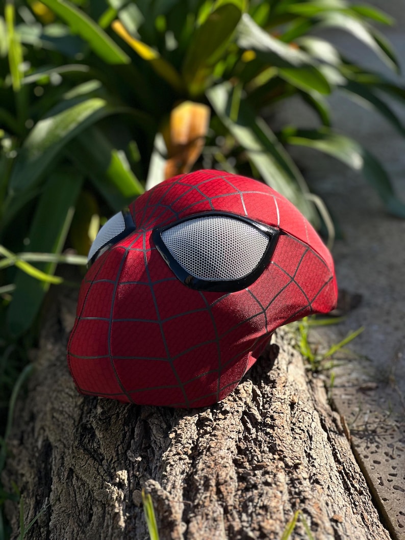 Puede incluir: Una m&aacute;scara roja de Spider-Man con telara&ntilde;as negras y lentes de malla blanca. La m&aacute;scara est&aacute; apoyada sobre un trozo de madera, con follaje verde en el fondo. Accesorio de disfraz de superh&eacute;roe.