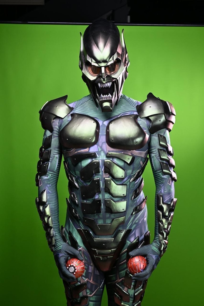 May include: A detailed costume of the Green Goblin, featuring a metallic helmet with menacing eyes and a jagged mouth. The suit has armored plating and is holding two red, spherical grenades. The background is a solid green.