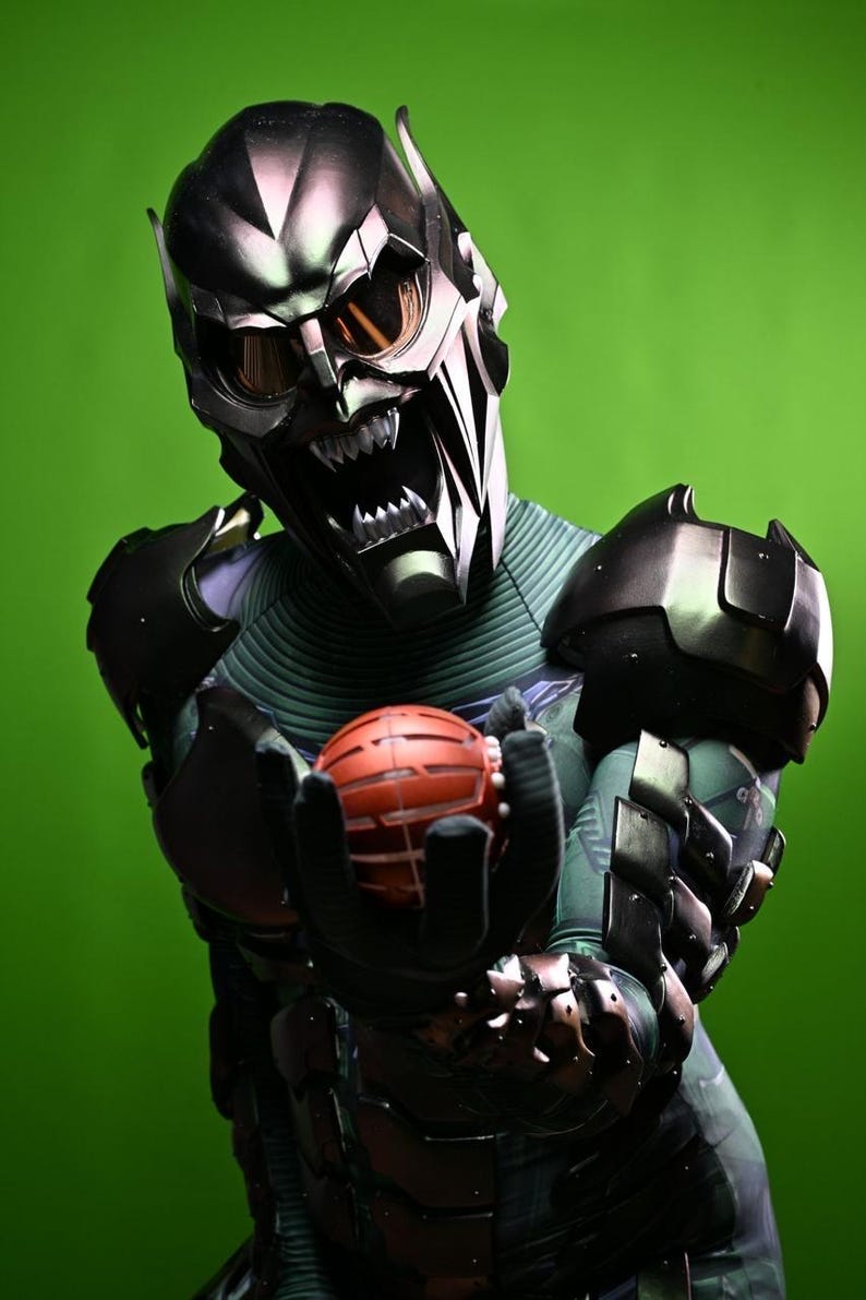 May include: A detailed image of a Green Goblin costume. The mask is metallic with sharp angles and menacing teeth. The costume features armored elements and a red, spherical grenade held in gloved hands. The background is a solid green.