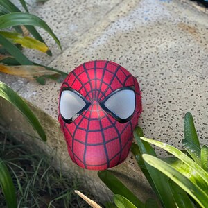 Może przedstawiać: Czerwono-czarna maska Spider-Mana z białymi siatkowymi osłonami oczu. Maska ma wz&oacute;r pajęczyny i spoczywa na betonowej powierzchni, otoczona zielonymi roślinami.