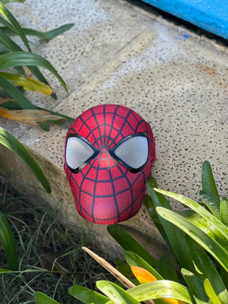 Może przedstawiać: Czerwono-czarna maska Spider-Mana z białymi siatkowymi osłonami oczu. Maska ma wz&oacute;r pajęczyny i spoczywa na betonowej powierzchni, otoczona zielonymi roślinami.