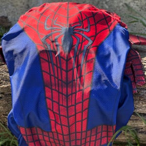 Może przedstawiać: Niebiesko-czerwony kostium Spider-Mana. Kostium ma czerwony wz&oacute;r pajęczyny na klatce piersiowej i plecach, z dużym czarnym emblematem pająka. Ramiona i rękawy są czerwone, a reszta kostiumu jest niebieska.