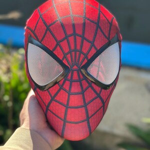 Może przedstawiać: Czerwono-czarna maska Spider-Mana z białymi siatkowymi osłonami oczu. Maska ma wz&oacute;r pajęczyny na czerwonej powierzchni i czarne kontury. Maska jest trzymana, prezentując wz&oacute;r i kształt.