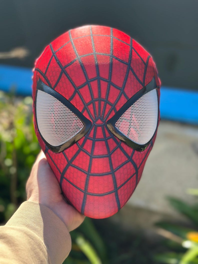 Może przedstawiać: Czerwono-czarna maska Spider-Mana z białymi siatkowymi osłonami oczu. Maska ma wz&oacute;r pajęczyny na czerwonej powierzchni i czarne kontury. Maska jest trzymana, prezentując wz&oacute;r i kształt.