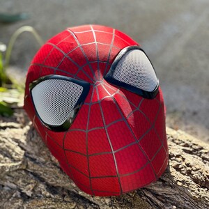 Puede incluir: Una m&aacute;scara roja de Spider-Man con un patr&oacute;n de telara&ntilde;a negro y cubiertas oculares de malla blanca. La m&aacute;scara est&aacute; sobre un trozo de madera, mostrando el dise&ntilde;o ic&oacute;nico.