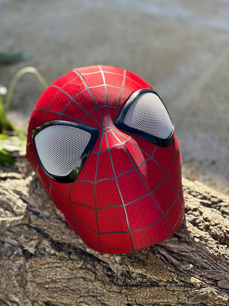 Puede incluir: Una m&aacute;scara roja de Spider-Man con un patr&oacute;n de telara&ntilde;a negro y cubiertas oculares de malla blanca. La m&aacute;scara est&aacute; sobre un trozo de madera, mostrando el dise&ntilde;o ic&oacute;nico.