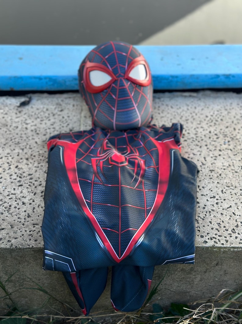 May include: A black and red Spider-Man costume with a mask featuring white eye lenses. The costume has a spider emblem on the chest and red web detailing. The suit is made of a textured fabric.