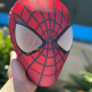 Może przedstawiać: Czerwona maska Spider-Mana z czarnym wzorem pajęczyny i białymi siatkowymi osłonami oczu. Maska jest trzymana, prezentując jej design i fakturę. Maska jest przedmiotem cosplay.