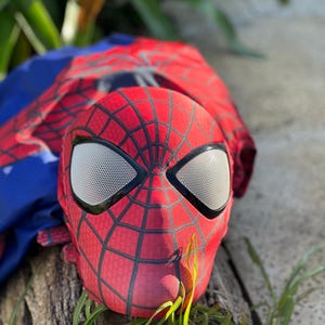 Może przedstawiać: Czerwono-czarna maska Spider-Mana z białymi siatkowymi osłonami oczu. Maska leży na kawałku drewna, z niebiesko-czerwonym kostiumem w tle. Zielona trawa na pierwszym planie.