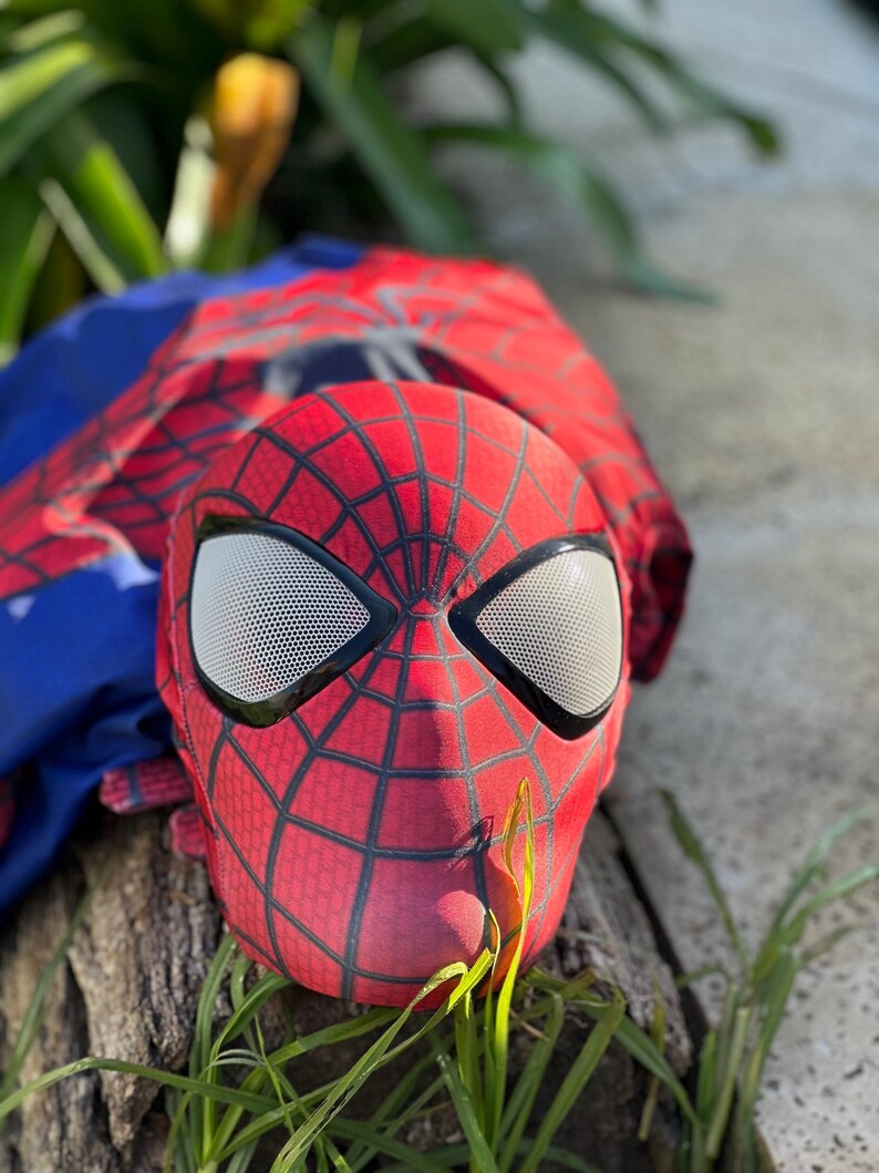 Może przedstawiać: Czerwono-czarna maska Spider-Mana z białymi siatkowymi osłonami oczu. Maska leży na kawałku drewna, z niebiesko-czerwonym kostiumem w tle. Zielona trawa na pierwszym planie.