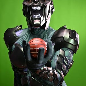 May include: A detailed costume of the Green Goblin, featuring a metallic mask with a menacing grin and sharp teeth. The costume includes a green and black armored suit, with the character holding a red pumpkin bomb.
