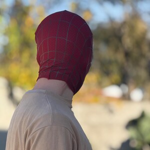 Puede incluir: Una m&aacute;scara de Spider-Man burdeos con un patr&oacute;n de telara&ntilde;a negra, usada por una persona. La m&aacute;scara cubre la cabeza y la cara, con la persona usando una camiseta beige de manga larga. El fondo est&aacute; borroso con tonos verdes y amarillos.