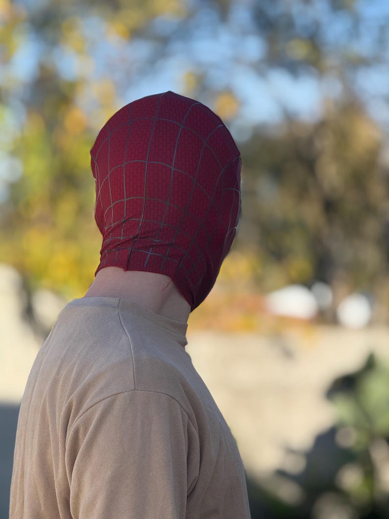 Puede incluir: Una m&aacute;scara de Spider-Man burdeos con un patr&oacute;n de telara&ntilde;a negra, usada por una persona. La m&aacute;scara cubre la cabeza y la cara, con la persona usando una camiseta beige de manga larga. El fondo est&aacute; borroso con tonos verdes y amarillos.