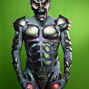 May include: A full-body costume featuring a metallic green and purple design. The costume includes a detailed helmet with a menacing expression and red-tinted lenses. The suit has armored plating and is holding two red, spherical objects.