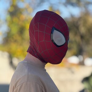 Puede incluir: Una m&aacute;scara de Spider-Man roja y negra con cubiertas oculares de malla blanca. La m&aacute;scara tiene un dise&ntilde;o de telara&ntilde;a. La persona que lleva la m&aacute;scara lleva una camiseta de manga larga de color beige. El fondo est&aacute; borroso con follaje verde y amarillo.