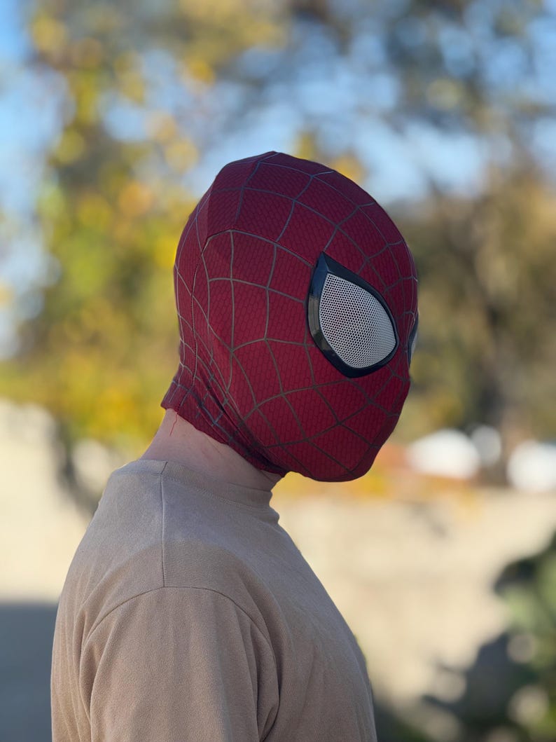 Puede incluir: Una m&aacute;scara de Spider-Man roja y negra con cubiertas oculares de malla blanca. La m&aacute;scara tiene un dise&ntilde;o de telara&ntilde;a. La persona que lleva la m&aacute;scara lleva una camiseta de manga larga de color beige. El fondo est&aacute; borroso con follaje verde y amarillo.