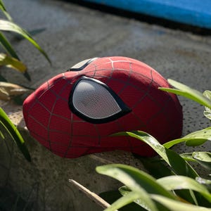 Puede incluir: Una m&aacute;scara roja de Spider-Man con telara&ntilde;as negras y cubiertas oculares de malla blanca. La m&aacute;scara est&aacute; hecha de una tela texturizada y es parcialmente visible, apoyada sobre una superficie de piedra.