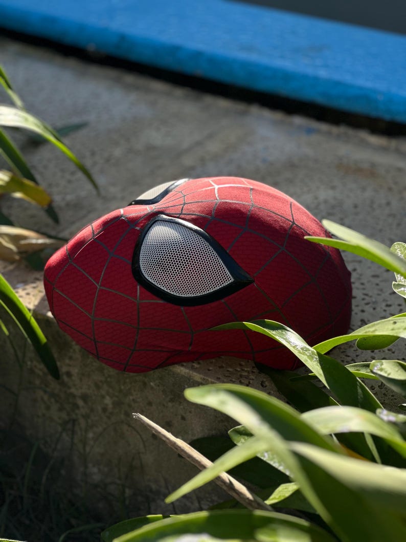 Puede incluir: Una m&aacute;scara roja de Spider-Man con telara&ntilde;as negras y cubiertas oculares de malla blanca. La m&aacute;scara est&aacute; hecha de una tela texturizada y es parcialmente visible, apoyada sobre una superficie de piedra.