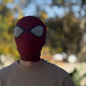 Puede incluir: Una m&aacute;scara de Spider-Man roja y negra con cubiertas oculares de malla blanca. La m&aacute;scara se usa sobre una sudadera beige. El fondo est&aacute; borroso con follaje verde y amarillo.