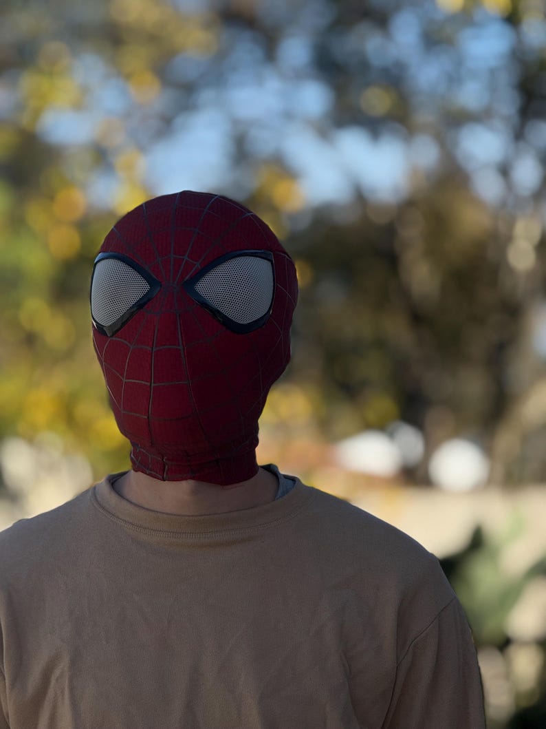 Puede incluir: Una m&aacute;scara de Spider-Man roja y negra con cubiertas oculares de malla blanca. La m&aacute;scara se usa sobre una sudadera beige. El fondo est&aacute; borroso con follaje verde y amarillo.