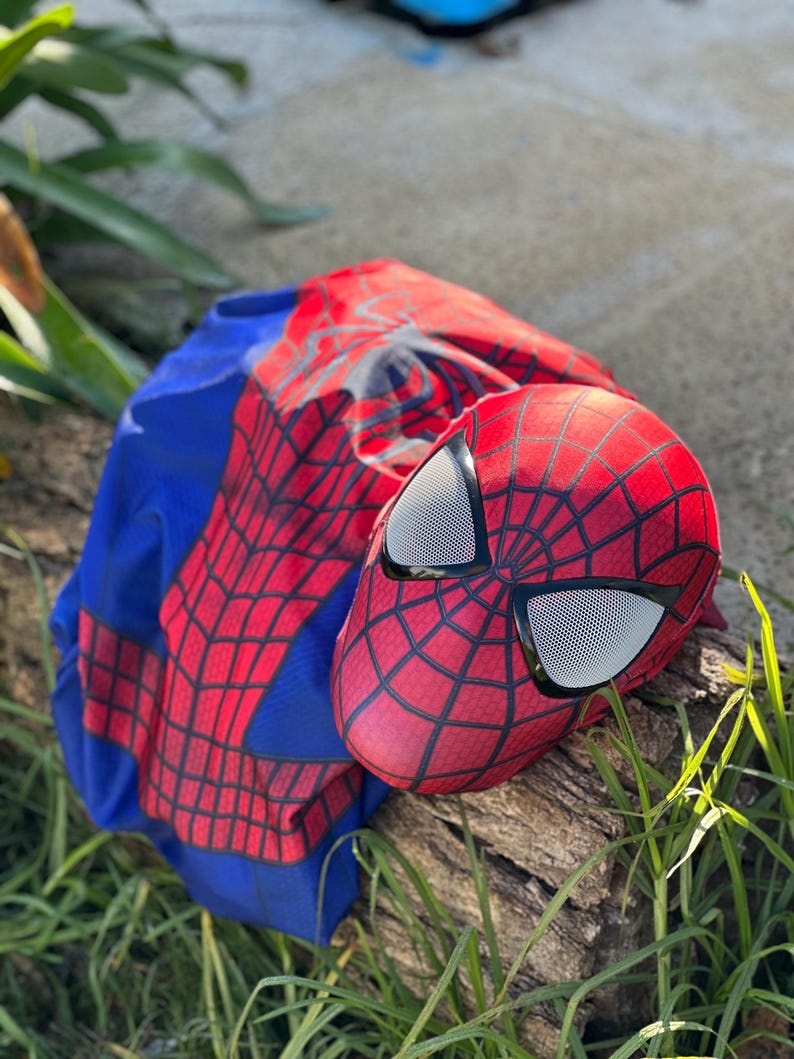 Może przedstawiać: Czerwono-czarna maska Spider-Mana z białymi siatkowymi osłonami oczu spoczywa na kłodzie obok niebiesko-czerwonego kostiumu. Maska ma wz&oacute;r pajęczyny i jest akcesorium do kostiumu superbohatera.