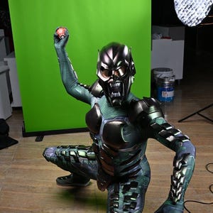 May include: A Green Goblin costume with a metallic mask and armor plating. The costume features a green and black color scheme with intricate details. The character is in a crouching position, holding a red object.