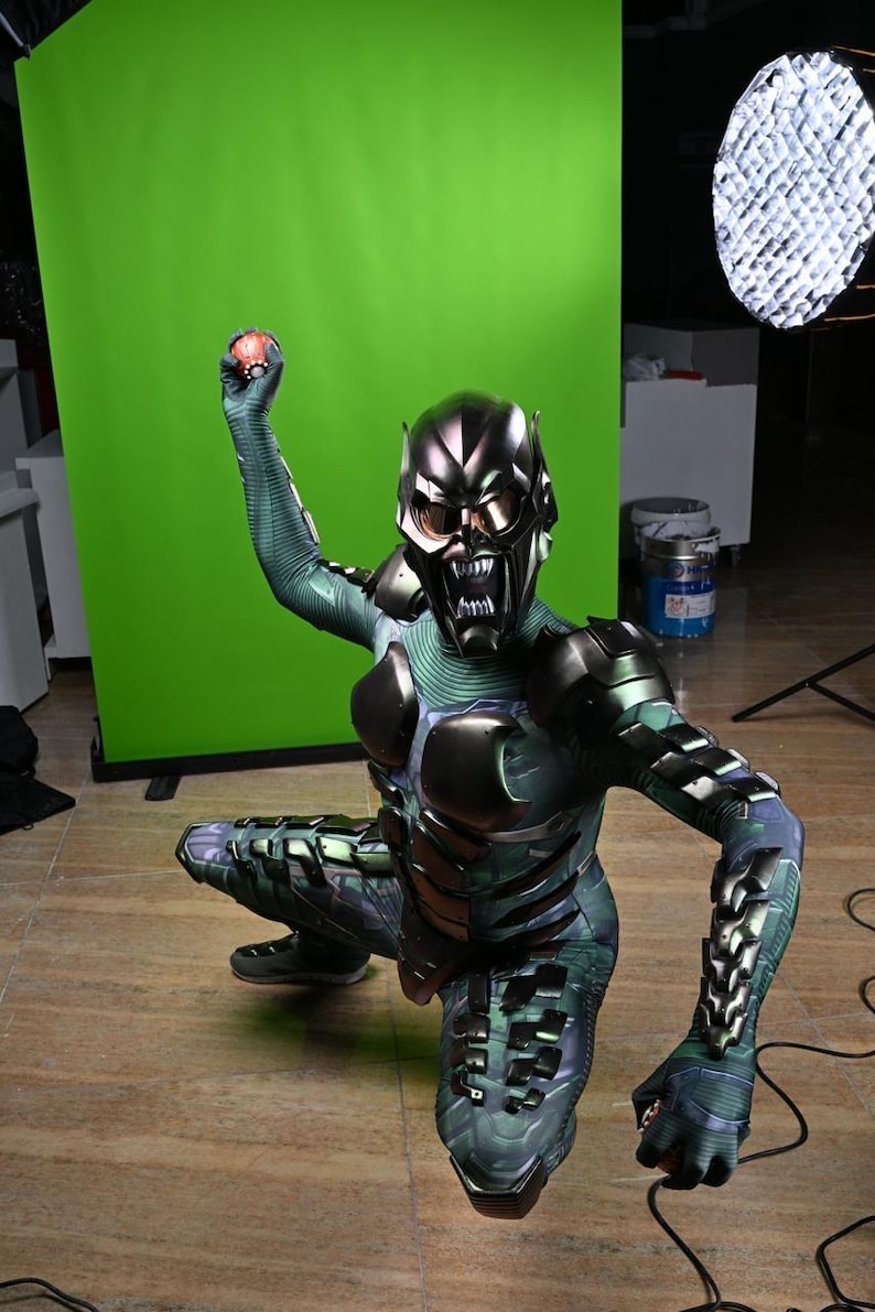 May include: A Green Goblin costume with a metallic mask and armor plating. The costume features a green and black color scheme with intricate details. The character is in a crouching position, holding a red object.