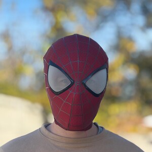 Puede incluir: Una m&aacute;scara de Spider-Man roja y negra con cubiertas oculares de malla blanca. La m&aacute;scara presenta un patr&oacute;n de telara&ntilde;a y se usa sobre una camiseta de color beige. El fondo est&aacute; borroso con tonos verdes y amarillos.