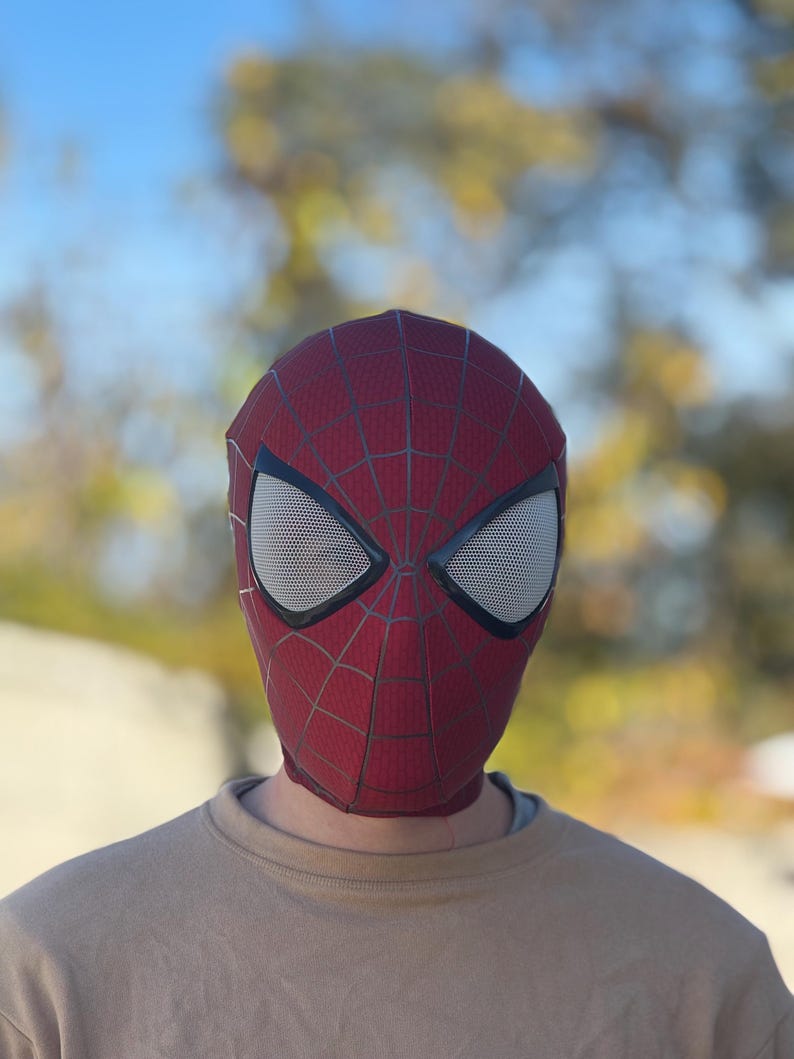 Puede incluir: Una m&aacute;scara de Spider-Man roja y negra con cubiertas oculares de malla blanca. La m&aacute;scara presenta un patr&oacute;n de telara&ntilde;a y se usa sobre una camiseta de color beige. El fondo est&aacute; borroso con tonos verdes y amarillos.