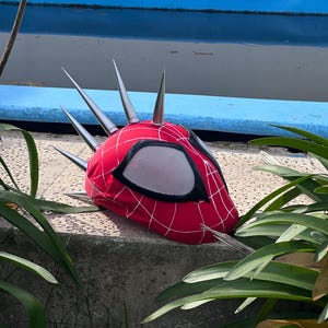 Op de afbeelding: Een rood Spider-Man-masker met een wit webpatroon en grote, zilveren spikes. Het masker heeft zwarte oogopeningen met een gaasmateriaal. Het masker ligt op een betonnen oppervlak met groene planten op de voorgrond.