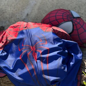 Może przedstawiać: Niebiesko-czerwony kostium Spider-Mana z pasującą maską. Kostium ma duży czerwony emblemat pająka na klatce piersiowej i czerwone wzory pajęczyny. Maska ma białe osłony oczu i czerwony wz&oacute;r pajęczyny.