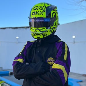 Puede incluir: Una persona con un casco de carreras verde neón con gráficos negros y una visera negra. La persona lleva una chaqueta negra con detalles morados y amarillos y un logotipo de Los Angeles Lakers.