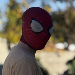 Puede incluir: Una m&aacute;scara de Spider-Man roja y negra con cubiertas oculares de malla blanca. La m&aacute;scara tiene un patr&oacute;n de telara&ntilde;a y se usa sobre la cabeza. La persona lleva una camiseta de manga larga de color beige.